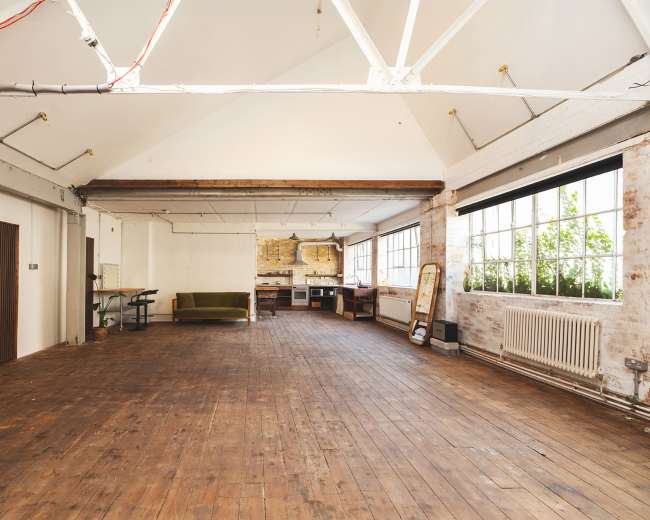 The image shows a spacious, open interior with wooden floors, large windows, and a simple kitchen area against one wall.