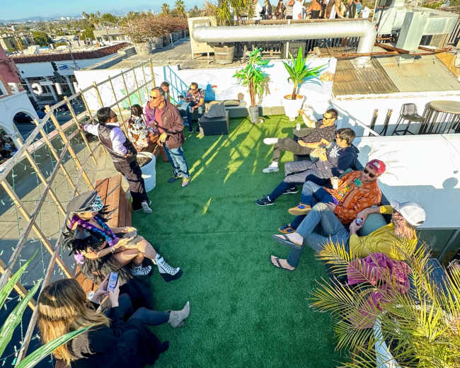 The image shows a rooftop gathering with people seated around on a green carpet under a clear blue sky.