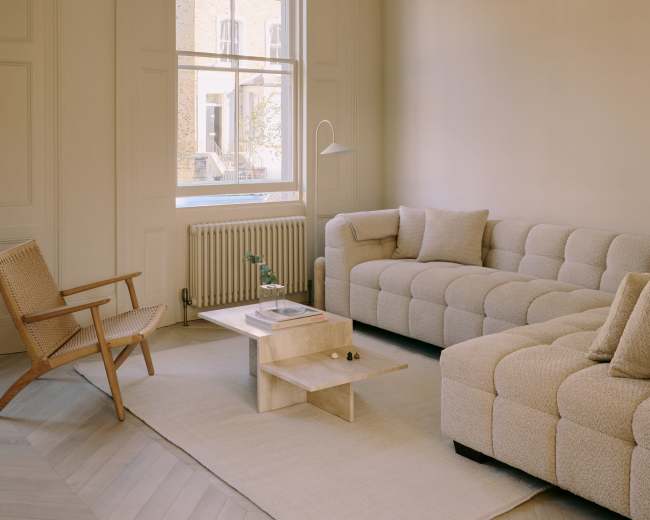 The image features a light-colored living room with a beige sectional sofa, a wooden armchair, a coffee table, and a large window allowing natural light to enter.