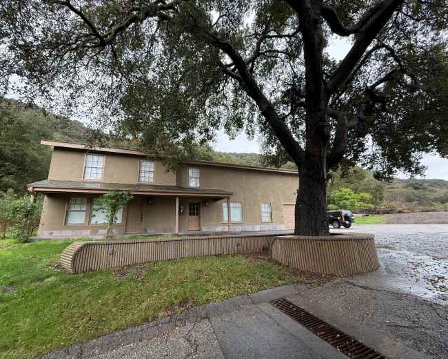 A two-story, beige building with a flat roof is set against a green landscape and a large tree on one side.