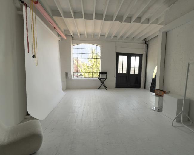 The image shows a spacious, well-lit interior with white walls, wooden beams, a black chair, and large windows.