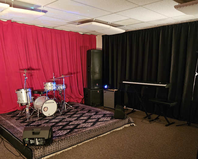 The image shows a music rehearsal space featuring a drum set, a keyboard, and a red curtain on one wall, with black curtains and carpet flooring.