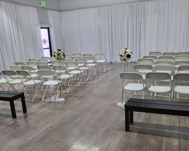 A spacious indoor venue set up for a ceremony, featuring rows of white folding chairs and flower arrangements on pedestals.