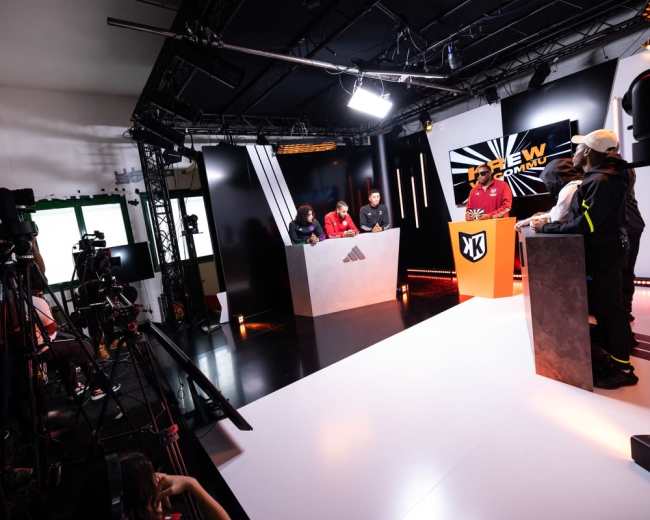 The image shows a modern studio setup with four participants at podiums and cameras positioned for a broadcast.