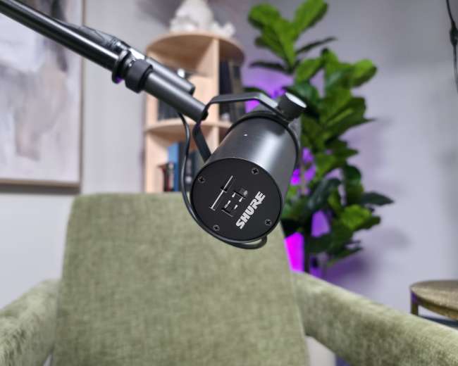 A close-up of a microphone mounted on a boom arm positioned near a green chair in a well-lit room with shelves and plants.