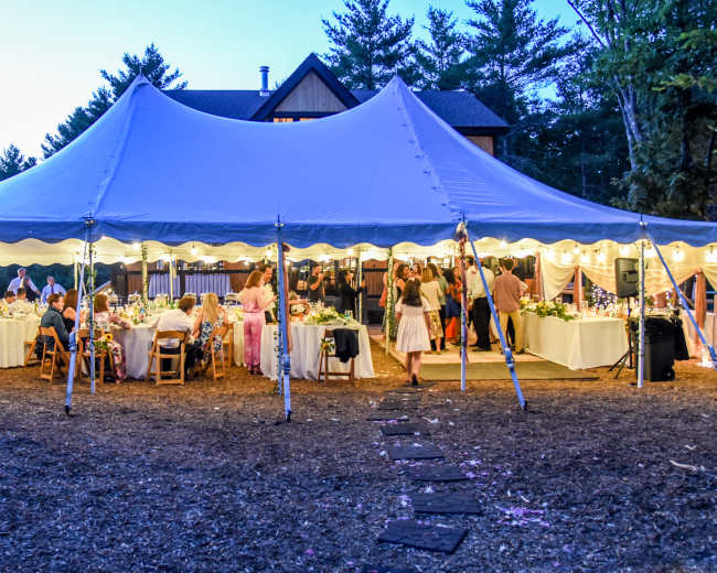 A large tent filled with tables and chairs is set up outdoors, where guests are gathered during an event near a house surrounded by trees.