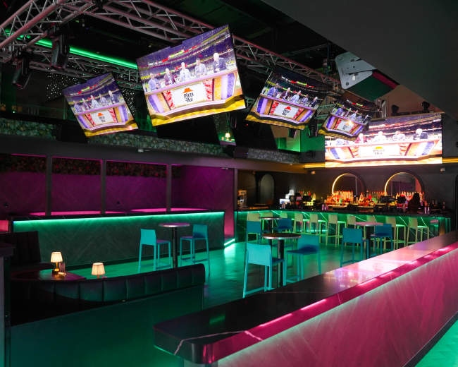 The image shows a modern bar interior with illuminated furniture and multiple screens displaying a sports event.