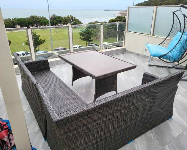 A wicker patio set with a table and two benches overlooks a grassy area and the ocean in the background.