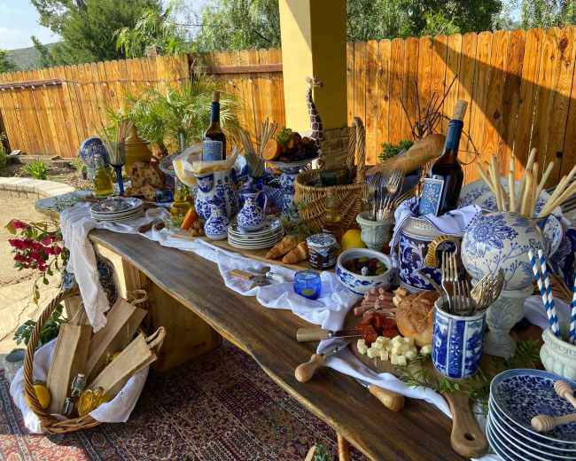 A wooden table is set with a variety of dishes, utensils, and decorative items, showcasing a spread of food and beverages accompanied by blue and white ceramic ware.