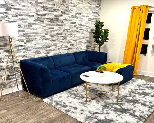 The image features a modern living room with a blue velvet sofa, a round white coffee table, a potted plant, and a yellow curtain against a stone-patterned wall.