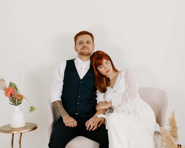 A couple sits closely together on a pink chair, with the woman resting her head on the man's shoulder in a softly decorated indoor setting.