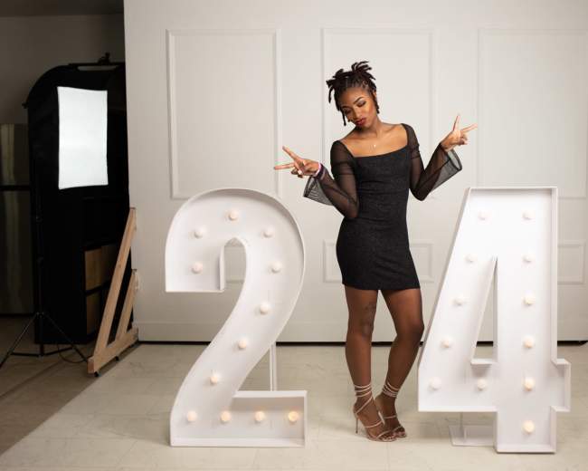 A woman poses between large illuminated numbers "24" while wearing a black dress in a well-lit studio setting.