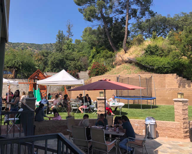 A sunny backyard gathering with people seated at tables under umbrellas, a trampoline, and a playground structure in the background.