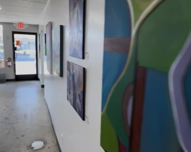 The image shows a corridor in an art gallery, featuring abstract paintings on the walls and a doorway leading outside at the far end.