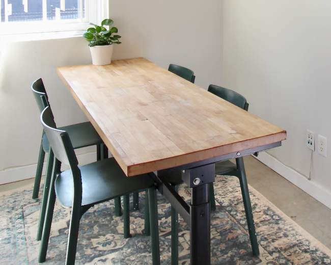 A wooden dining table with four green chairs is positioned in a corner of a room with light-colored walls and a window, featuring a small potted plant and artwork on the wall.