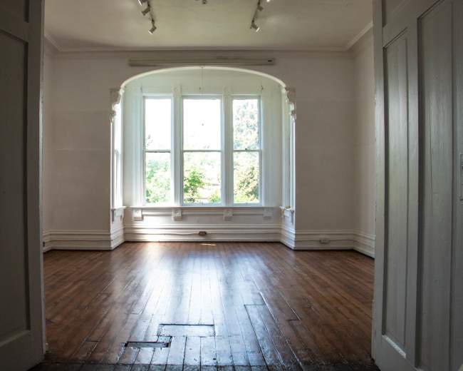 A spacious, well-lit room with wooden floors and large windows facing a green outside view.