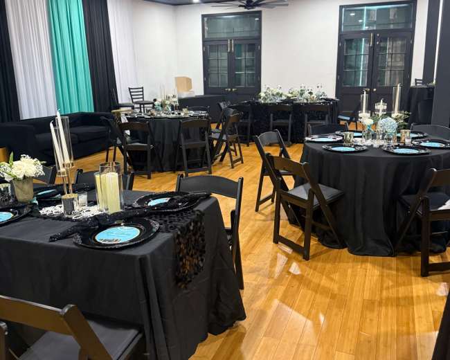 The image shows a banquet hall arranged with black tablecloths, turquoise centerpieces, and a wooden floor, featuring several tables and chairs.
