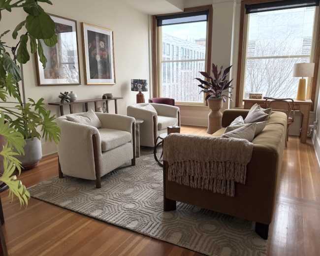 The image shows a cozy interior living room with two armchairs, a sofa, decorative art on the walls, and large windows letting in natural light.