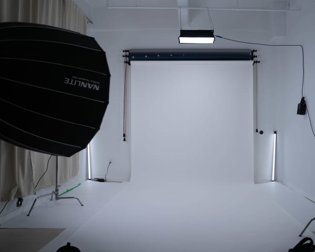 The image shows a photography studio with a white backdrop, softbox lighting, and various equipment along the walls.