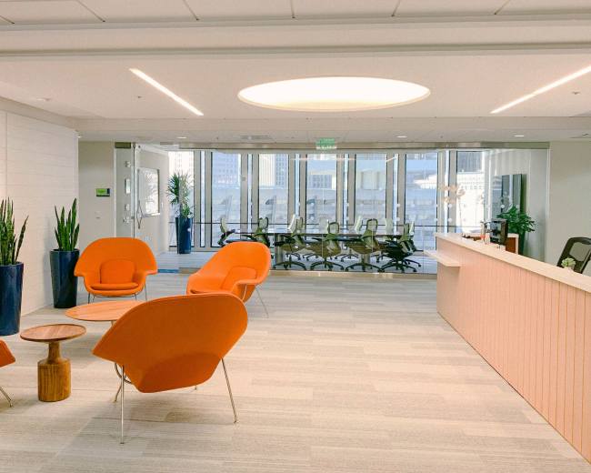 The image depicts a modern office lobby featuring orange chairs, a wooden side table, large indoor plants, and a glass conference room with a meeting table in the background.