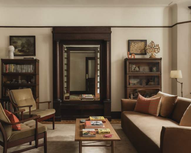 The image shows a cozy living room featuring a brown sofa, wooden bookshelves, a mirror, and a coffee table with various items on it.