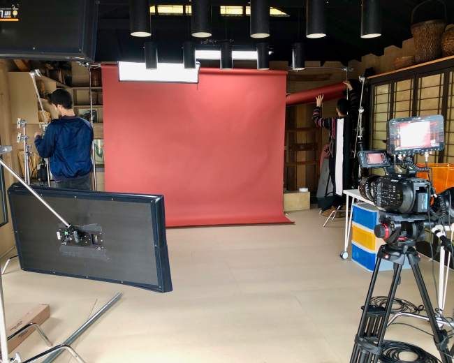 A photography studio features a red backdrop, lighting equipment, and a camera setup, with two people adjusting the scene.