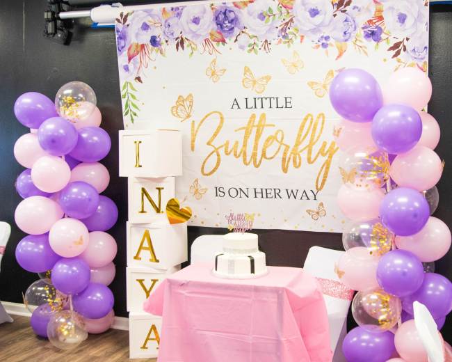 A decorated party space features a banner with floral designs, a cake on a table, and balloon arrangements in pink, purple, and gold.