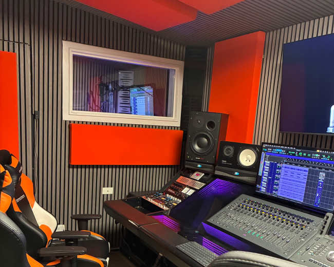 The image shows a music production studio with a soundboard, two gaming-style chairs, and soundproofing panels on the walls.