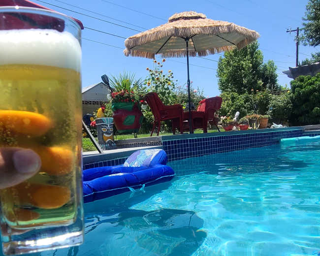 A hand holds a glass of beer above a clear swimming pool with a floating lounge and a shaded seating area in the background.