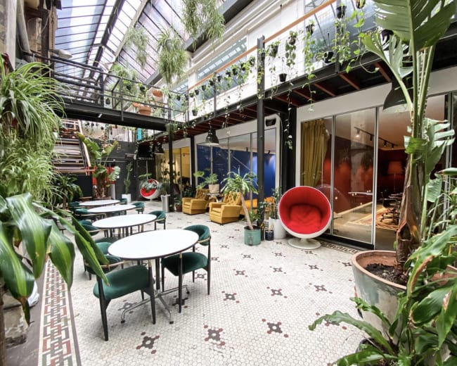 A modern indoor space filled with various plants, featuring green chairs and tables, large windows, and vibrant decor.