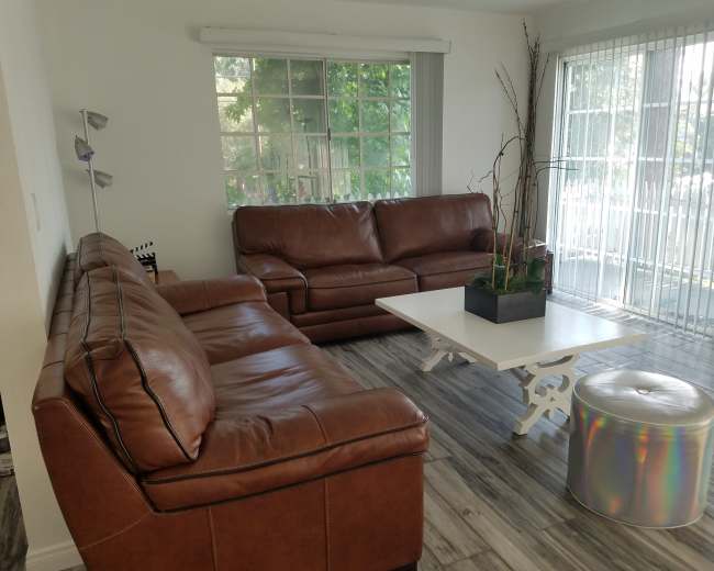 The image shows a living room with two brown leather sofas, a white coffee table, and a metallic stool near a large window with natural light.