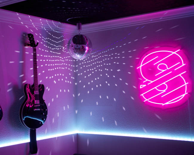 The image features two electric guitars hanging on a wall with a disco ball above and vibrant colored lights creating patterns on the ceiling.
