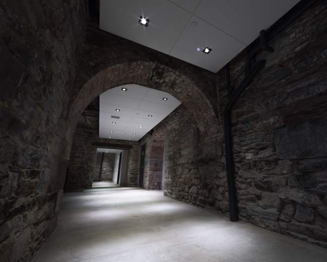 The image shows a dimly lit corridor with exposed stone walls and a ceiling that features modern lighting fixtures.