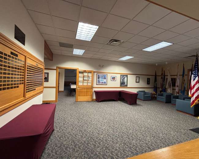 The interior of a room features a carpeted floor, two maroon tables, blue seating, several flags, and walls adorned with photographs.