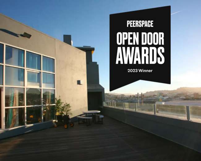 A modern rooftop space with large windows, a wooden deck, and a table set for outdoor seating, highlighted by a banner announcing the Peerspace Open Door Awards 2023 Winner.