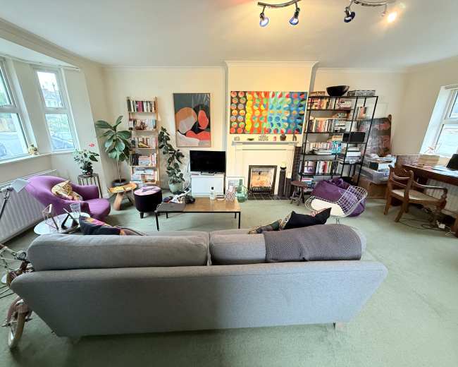 The image shows a cozy living room with a gray sofa, two purple armchairs, a television, shelves filled with books, and colorful artwork on the walls.