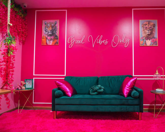 The image shows a vibrant pink room featuring a green couch with decorative pillows, framed animal portraits on the walls, and the phrase "Good Vibes Only" written in cursive.