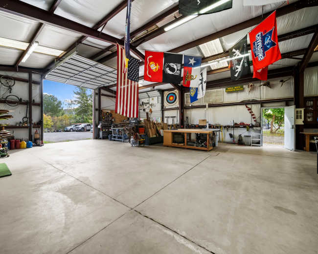 The image shows an expansive industrial garage or workshop with metal framing, concrete flooring, and various tools and equipment arranged along the walls, along with multiple flags hanging from the ceiling.