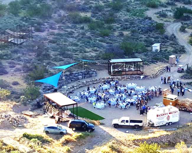 An outdoor gathering is set up in a cleared area with tables and chairs, surrounded by desert vegetation and structures in the background.