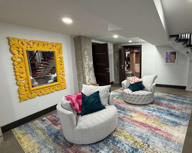The image shows a modern basement area featuring two upholstered chairs on a colorful striped rug, framed by exposed concrete columns and bright wall decor.