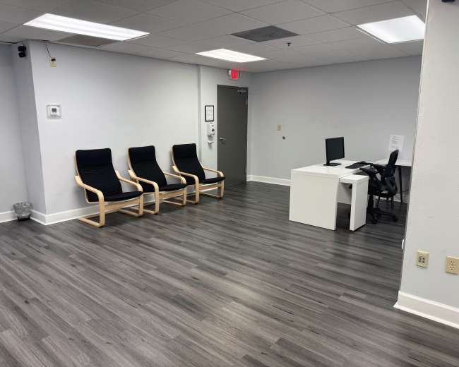 The image shows a modern waiting room with three chairs, a small desk with a computer, and a printer in the corner.