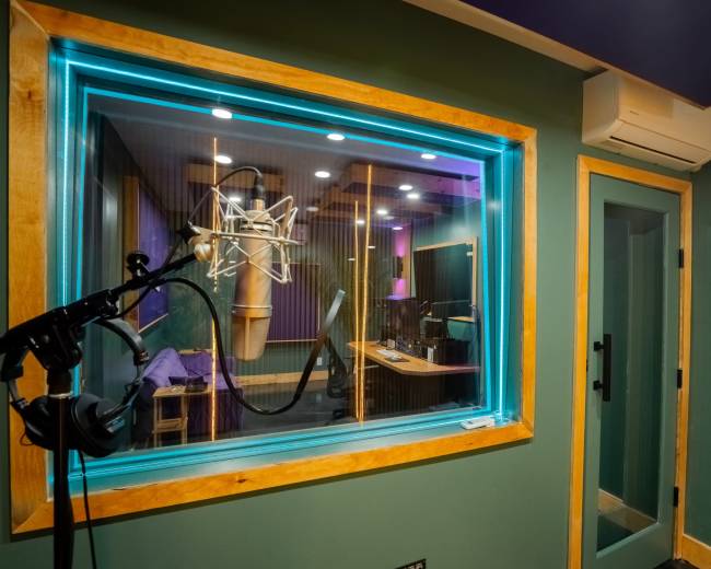 The image shows a soundproof recording studio with a microphone set up and a view into another room featuring sound equipment and a computer.