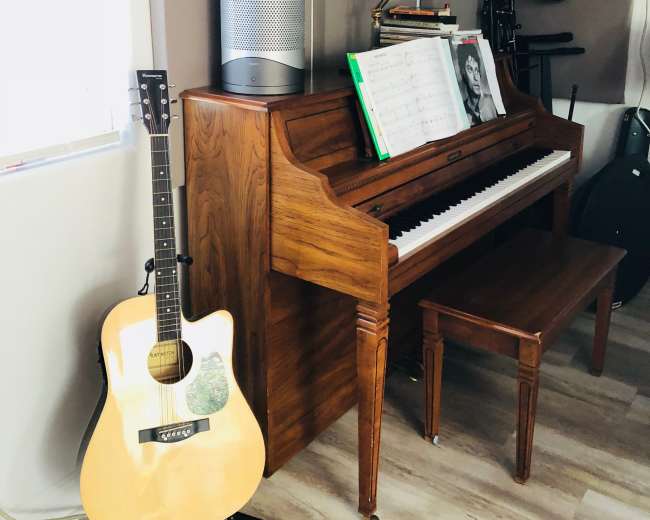 A wooden piano with sheet music sits beside an acoustic guitar in a well-lit music room.