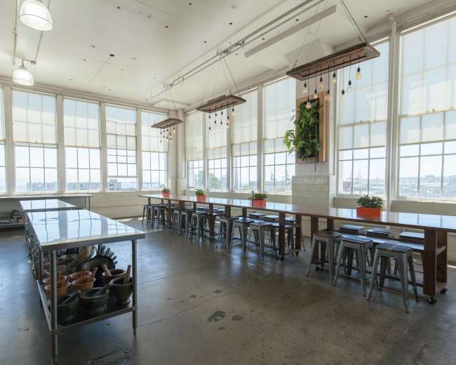 A spacious kitchen with large windows, featuring long communal tables, metal stools, and potted plants along the walls.
