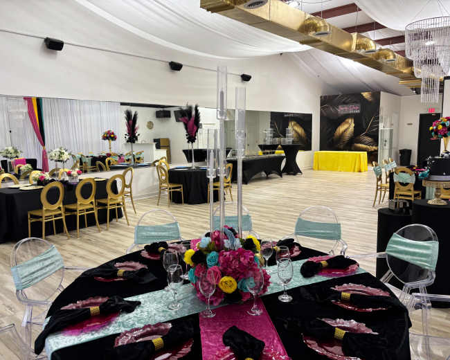 The image shows a brightly decorated event space with elegantly set tables featuring colorful centerpieces and a mix of black and vibrant tablecloths, along with a mirrored wall and draped fabric overhead.