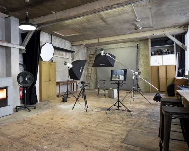 A photography studio features multiple lighting setups, a backdrop, and wooden flooring, with equipment strategically arranged around the space.