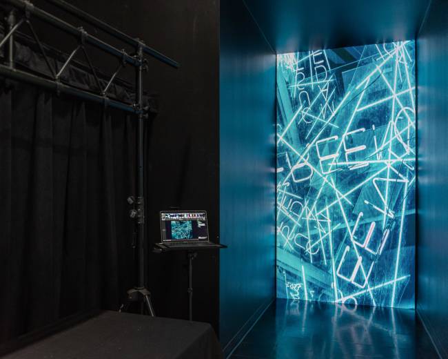 The image shows a darkened corridor leading to a brightly lit wall displaying blue digital text, with a black curtain and a laptop on a stand nearby.