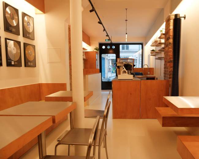 The image shows the interior of a modern café with wooden furniture, a counter at the rear, and framed circular plaques on the walls.
