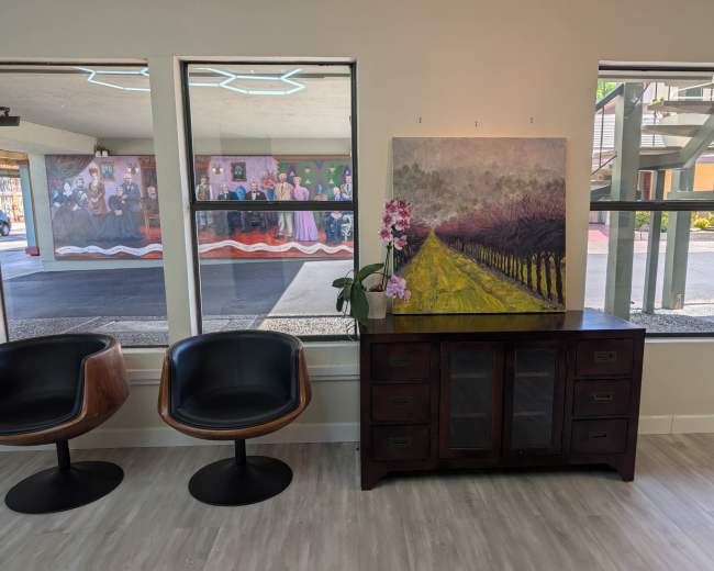 The image shows a modern interior with two black chairs, a wooden sideboard displaying a large painting of a vineyard, and windows revealing a mural and outdoor area.