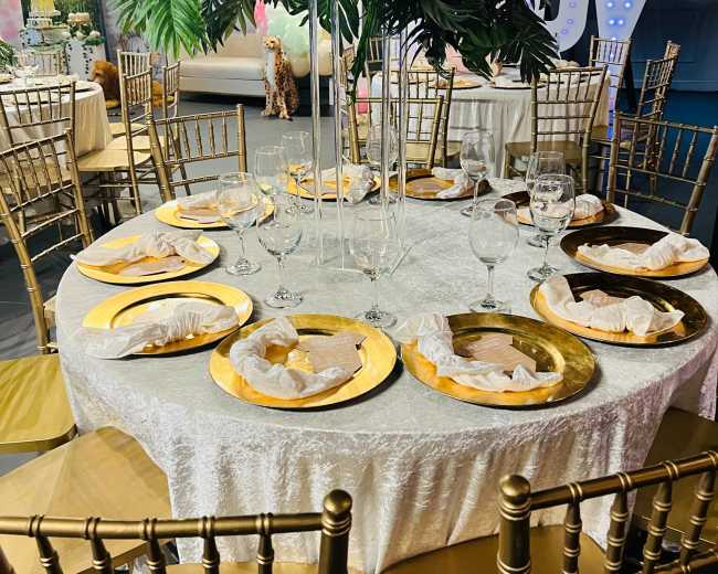 A beautifully arranged dining setup features round tables covered with white tablecloths, each adorned with gold chargers, glasses, and decorative centerpiece plants.
